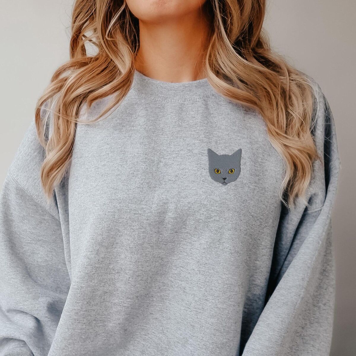 Grey Cat Sweatshirt Sport Grey Woman with long wavy hair in a Sport Grey crewneck sweatshirt with a gray cat face with gold eyes embroidered on the left chest