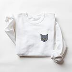 White crewneck sweatshirt laid flat on a white background with a gray cat face with gold eyes embroidered on the left chest