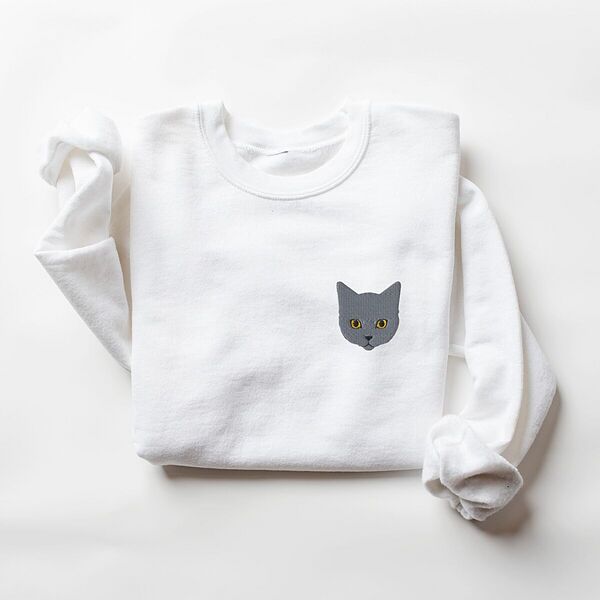 White crewneck sweatshirt laid flat on a white background with a gray cat face with gold eyes embroidered on the left chest