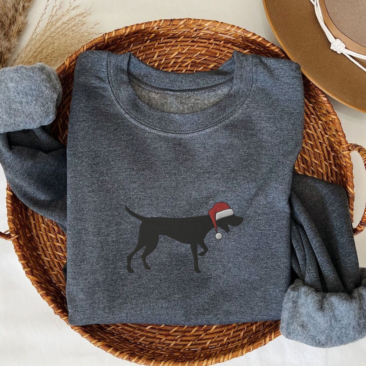 GSP Christmas Sweatshirt – Dark Heather Dark Heather crewneck sweatshirt folded in a wicker basket, embroidered with a black German shorthaired pointer silhouette in a red Santa hat