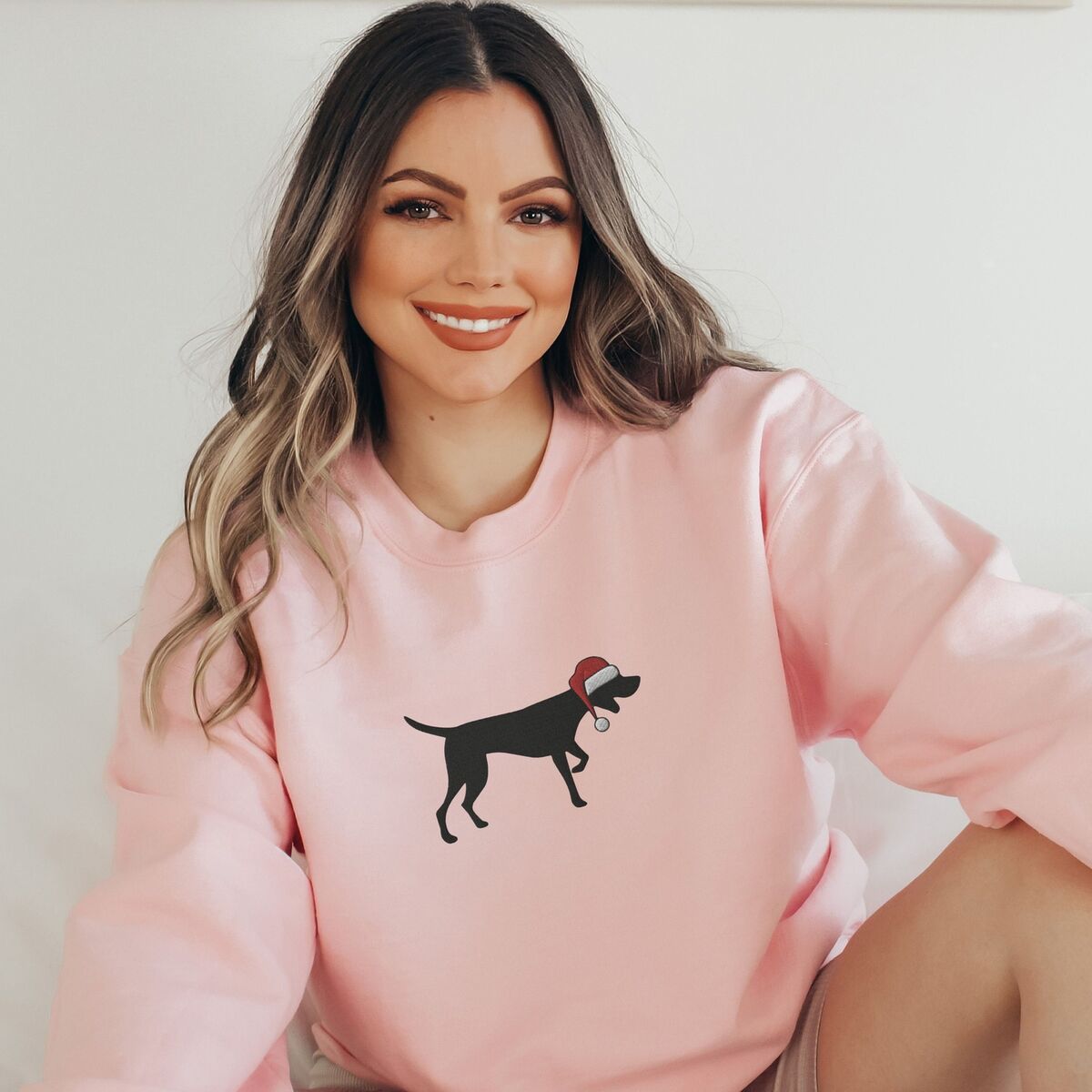 GSP Christmas Sweatshirt - Light Pink Smiling woman sitting in a Light Pink crewneck sweatshirt embroidered with a black German shorthaired pointer silhouette in a red Santa hat