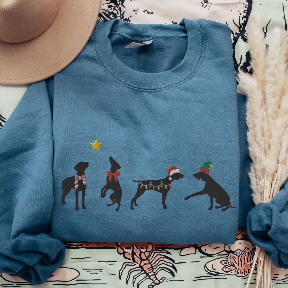 GSP Silhouette Christmas Sweatshirt – Indigo Blue Indigo Blue crewneck sweatshirt laid flat with a tan hat and dried pampas grass, embroidered with four black German shorthaired pointer silhouettes in a striped scarf, a red bow, Christmas lights, a Santa hat and an elf hat under a gold star