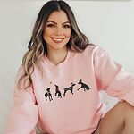 Smiling woman sitting in a Light Pink crewneck sweatshirt embroidered with four black German shorthaired pointer silhouettes in a striped scarf, a red bow, Christmas lights, a Santa hat and an elf hat under a gold star