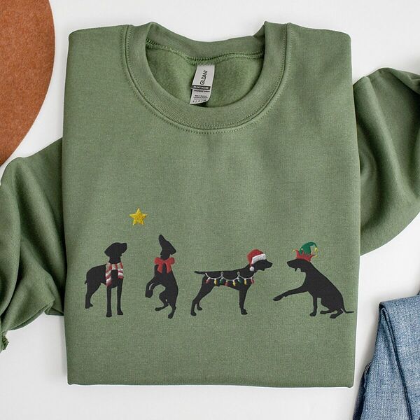 Military Green crewneck sweatshirt laid flat beside jeans, embroidered with four black German shorthaired pointer silhouettes in a striped scarf, a red bow, Christmas lights, a Santa hat and an elf hat under a gold star