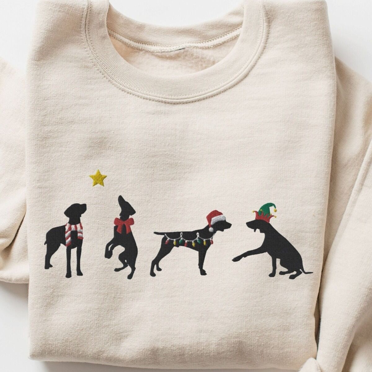 GSP Silhouette Christmas Sweatshirt – Sand Sand crewneck sweatshirt laid flat on a white background, embroidered with four black German shorthaired pointer silhouettes in a striped scarf, a red bow, Christmas lights, a Santa hat and an elf hat under a gold star