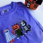 Close-up of the embroidered ghost cat, pumpkin cat and skeleton cat on a violet tee