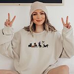 Smiling woman in a beanie making peace signs in a sand crewneck sweatshirt, embroidered with four black pit bull silhouettes with a jack-o'-lantern, an orange bandana, a purple witch hat and bats
