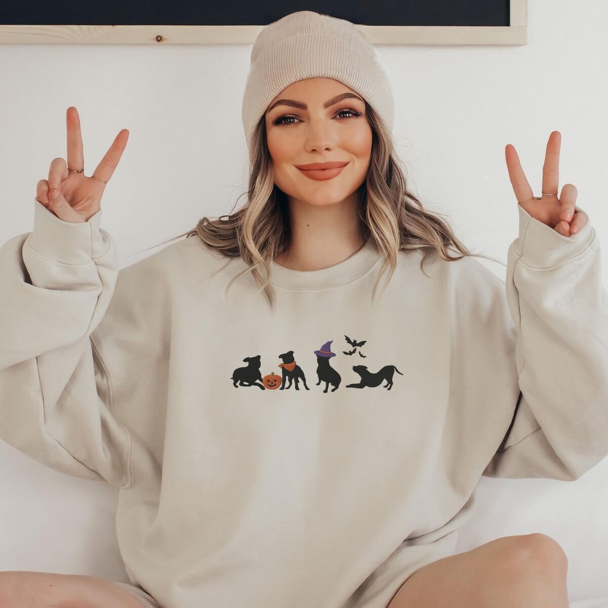 Halloween Pitbull Sweatshirt - worn Smiling woman in a beanie making peace signs in a sand crewneck sweatshirt, embroidered with four black pit bull silhouettes with a jack-o'-lantern, an orange bandana, a purple witch hat and bats