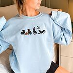 Woman with her hair up in a Light Blue crewneck sweatshirt, embroidered with four black pit bull silhouettes with a jack-o'-lantern, an orange bandana, a purple witch hat and bats