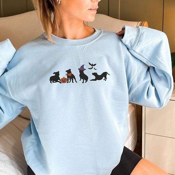 Woman with her hair up in a Light Blue crewneck sweatshirt, embroidered with four black pit bull silhouettes with a jack-o'-lantern, an orange bandana, a purple witch hat and bats