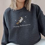 Woman in a Dark Heather crewneck sweatshirt embroidered with a ghost dachshund on a broomstick among stars and the words Happy Halloweenie