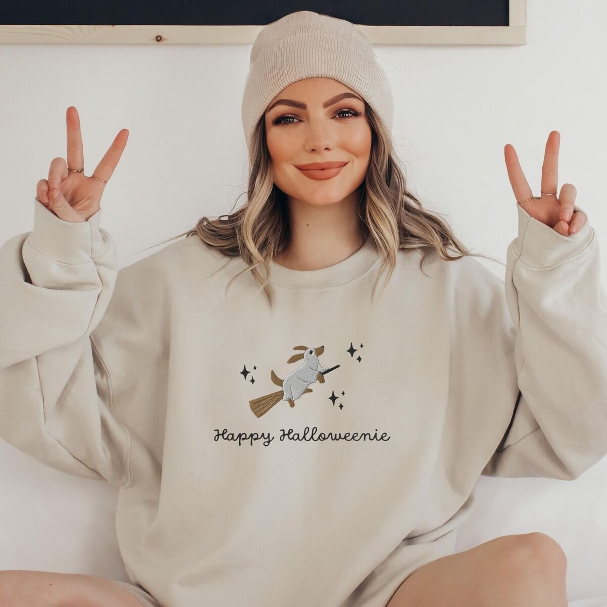 Happy Halloweenie Sweatshirt - worn Woman in a beanie making peace signs in a Sand crewneck sweatshirt embroidered with a ghost dachshund on a broom and the words Happy Halloweenie