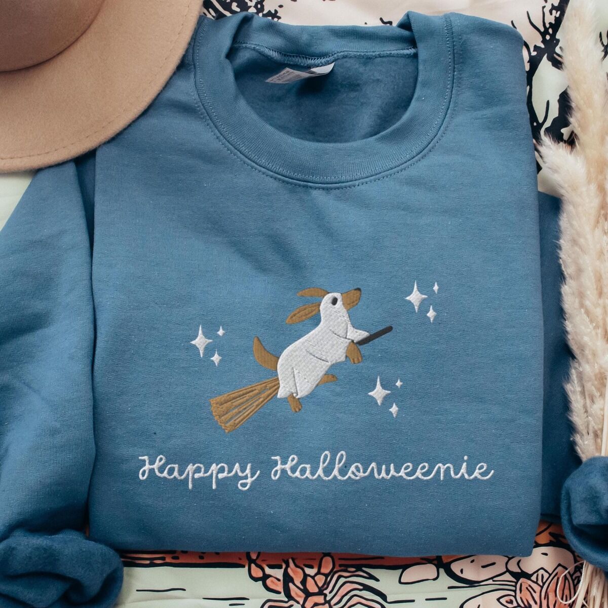 Happy Halloweenie Sweatshirt – Indigo Blue Indigo Blue crewneck sweatshirt laid flat with a felt hat, embroidered with a ghost dachshund flying on a broomstick among white stars above the words Happy Halloweenie