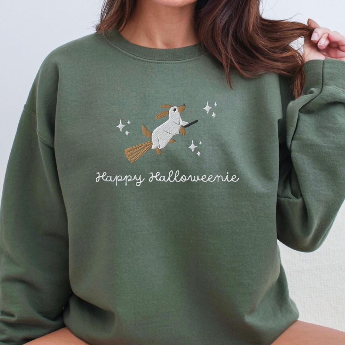 Happy Halloweenie Sweatshirt – Military Green Woman in a Military Green crewneck sweatshirt embroidered with a ghost dachshund on a broom and the words Happy Halloweenie in white script