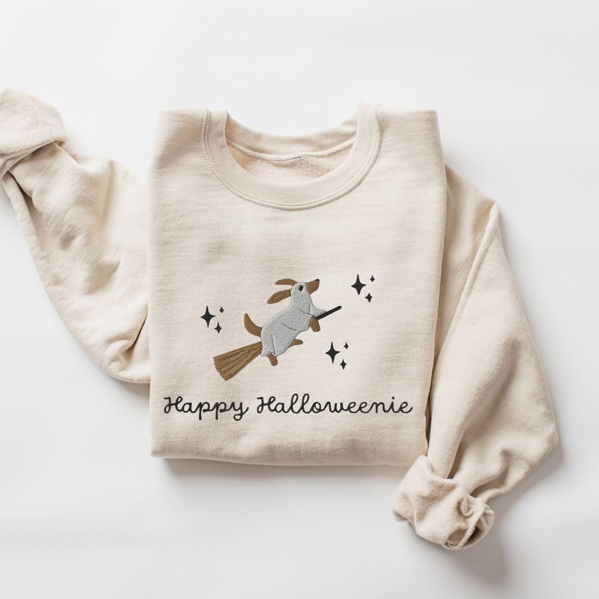 Happy Halloweenie Sweatshirt – Sand Sand crewneck sweatshirt laid flat on white, embroidered with a dachshund in a ghost sheet riding a broom among stars above the words Happy Halloweenie in black script