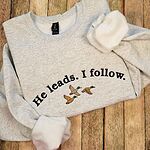 Ash Gildan crewneck sweatshirt folded on wooden boards with the cuffs turned up, embroidered with the arched black words He leads. I follow. above three mallard ducks in flight