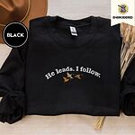 Black crewneck sweatshirt with a brown felt hat and dried bunny tail grass, embroidered with three mallard ducks flying under the arched words He leads. I follow. in white thread