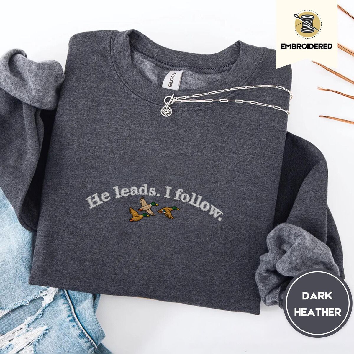 He Leads I Follow Sweatshirt – Dark Heather Dark Heather crewneck sweatshirt folded with ripped jeans and a silver chain necklace, embroidered with three mallard ducks flying under the arched words He leads. I follow. in white thread