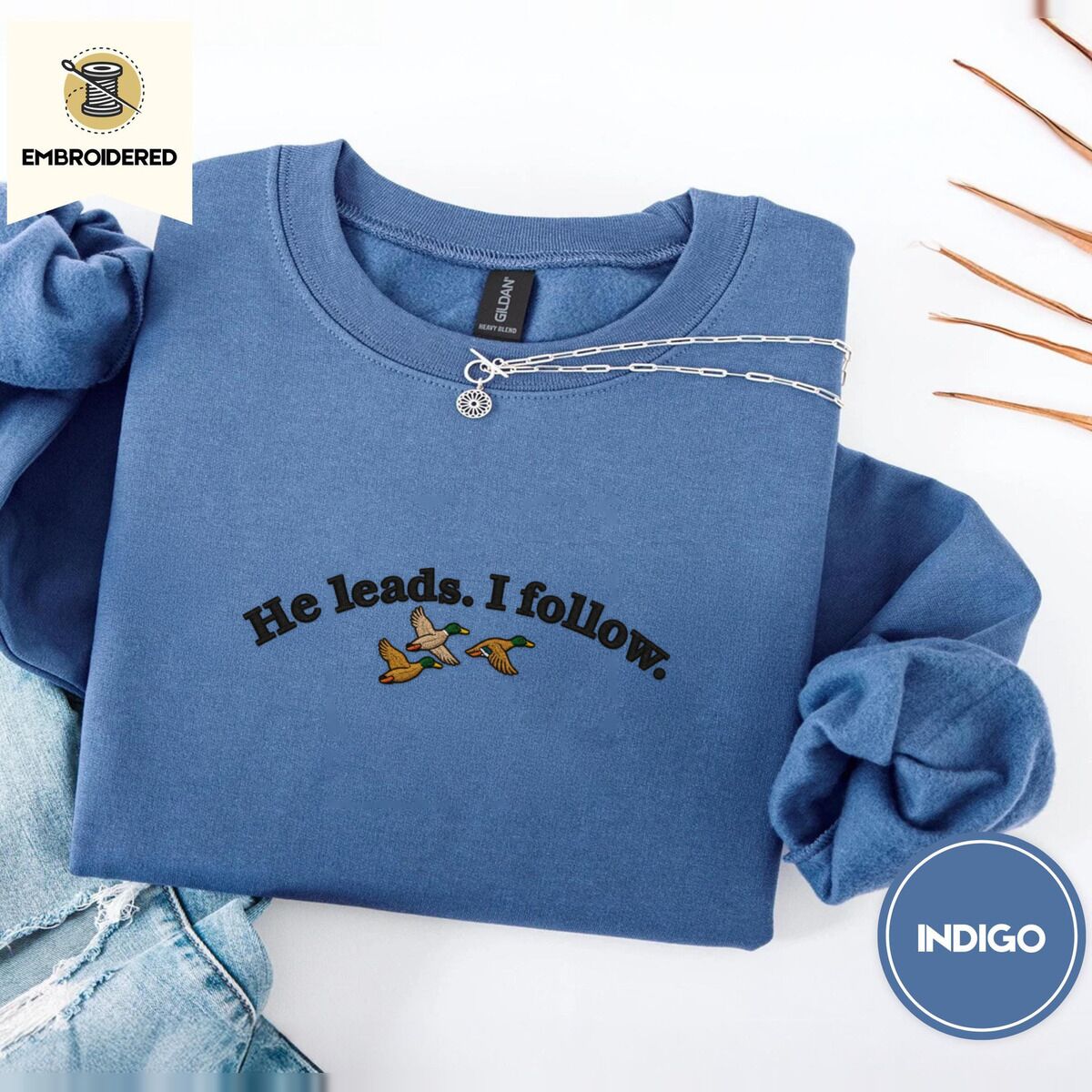 He Leads I Follow Sweatshirt – Indigo Blue Indigo Blue crewneck sweatshirt folded with light jeans and dried twigs, embroidered with three mallard ducks flying under the arched words He leads. I follow. in black thread