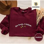 Maroon crewneck sweatshirt with a brown felt hat and dried bunny tail grass, embroidered with three mallard ducks flying under the arched words He leads. I follow. in white thread