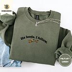 Military Green crewneck sweatshirt folded with light jeans and a silver chain necklace, embroidered with three mallard ducks flying under the arched words He leads. I follow. in black thread