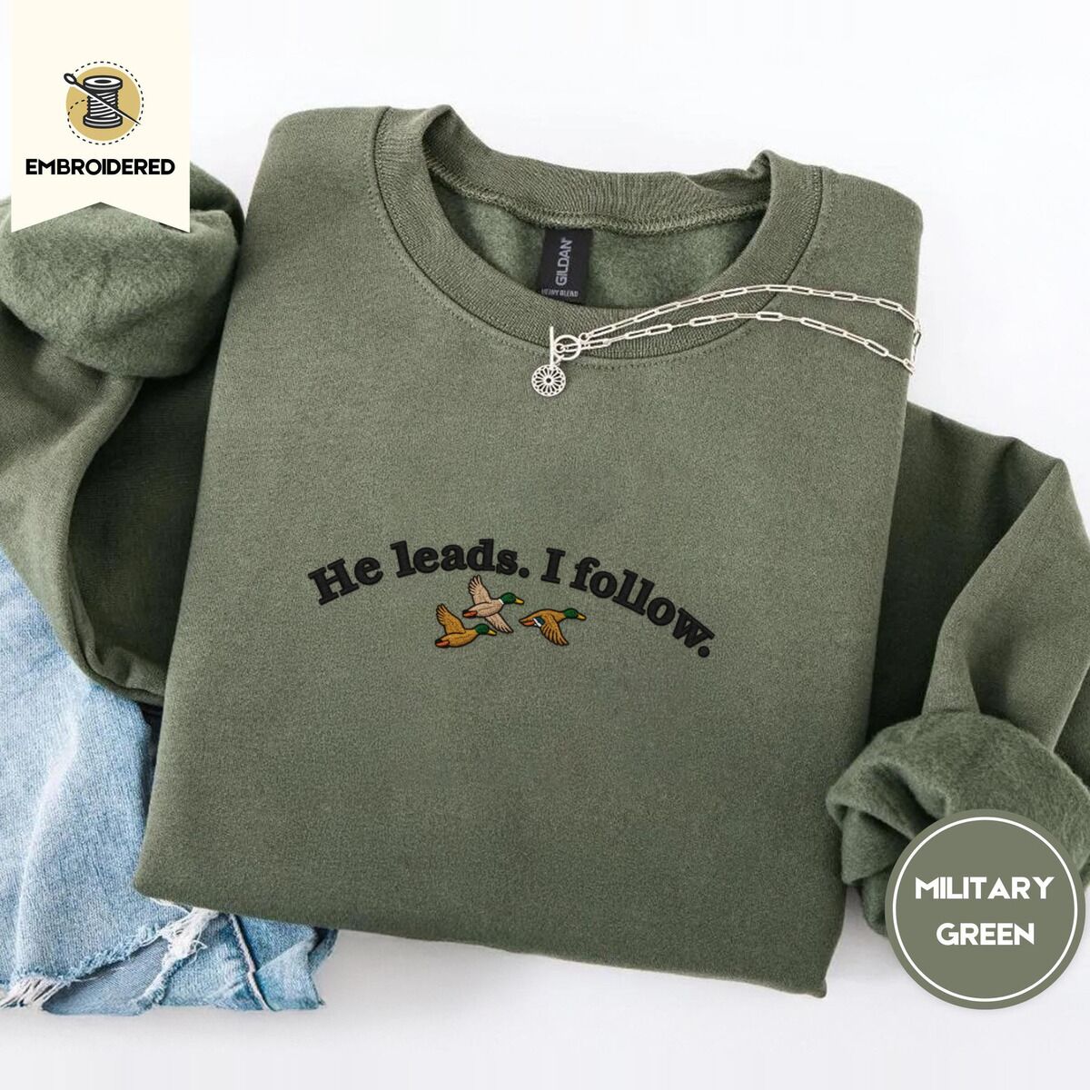 He Leads I Follow Sweatshirt – Military Green Military Green crewneck sweatshirt folded with light jeans and a silver chain necklace, embroidered with three mallard ducks flying under the arched words He leads. I follow. in black thread