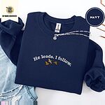 Navy crewneck sweatshirt folded with ripped jeans and a silver chain necklace, embroidered with three mallard ducks flying under the arched words He leads. I follow. in white thread