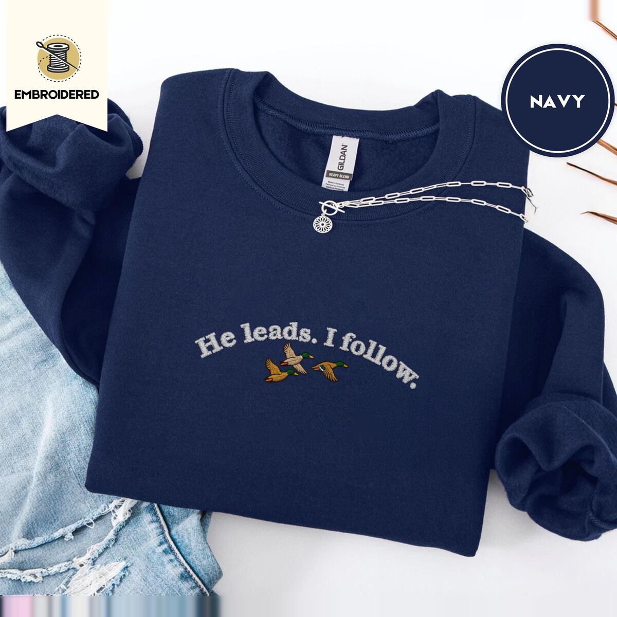 He Leads I Follow Sweatshirt – Navy Navy crewneck sweatshirt folded with ripped jeans and a silver chain necklace, embroidered with three mallard ducks flying under the arched words He leads. I follow. in white thread