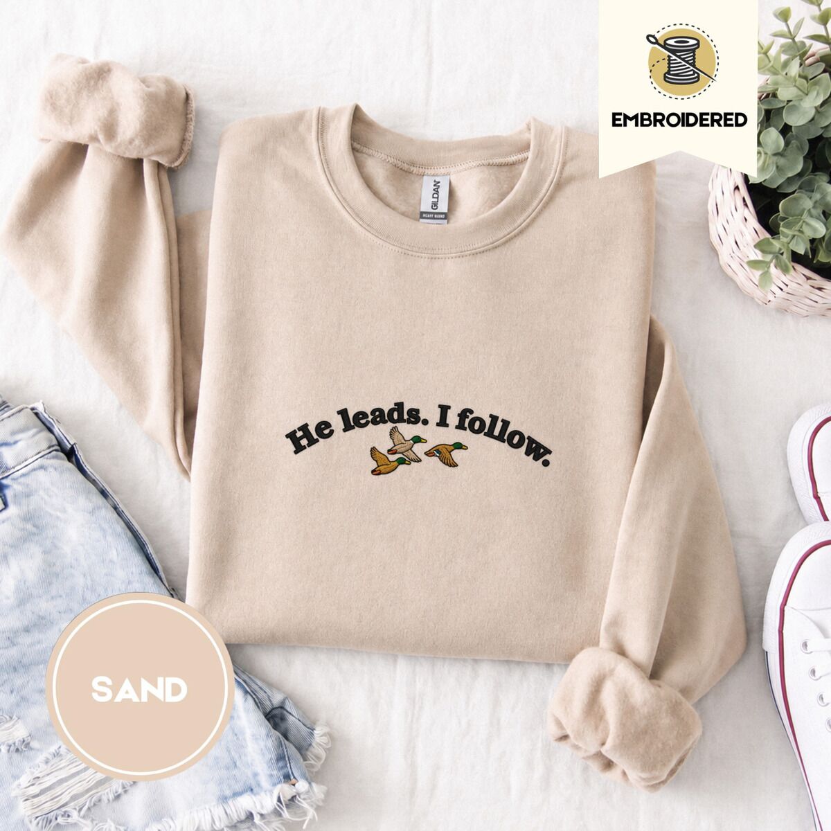 He Leads I Follow Sweatshirt – Sand Sand crewneck sweatshirt folded with denim shorts, white sneakers and a potted plant, embroidered with three mallard ducks flying under the arched words He leads. I follow. in black thread
