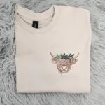 Highland cow Christmas sweatshirt in Sand with a shaggy embroidered highland cow wearing holly and pine on the left chest