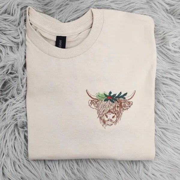 Highland cow Christmas sweatshirt in Sand with a shaggy embroidered highland cow wearing holly and pine on the left chest