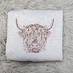 Ash crewneck sweatshirt folded on a gray faux fur rug, embroidered in brown thread with a shaggy Highland cow head with long curved horns