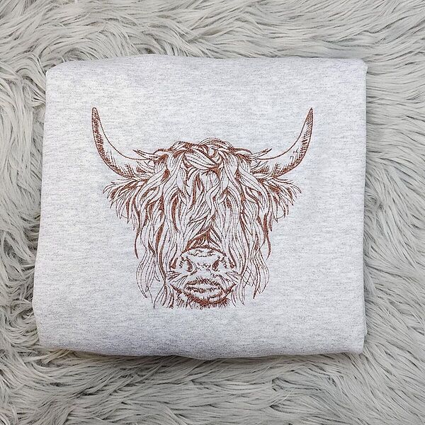 Ash crewneck sweatshirt folded on a gray faux fur rug, embroidered in brown thread with a shaggy Highland cow head with long curved horns