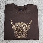 Dark Chocolate crewneck sweatshirt folded on a gray faux fur rug, embroidered in light tan thread with a shaggy Highland cow head with long curved horns