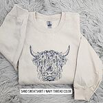 Sand crewneck sweatshirt on a gray faux fur rug with the shaggy Highland cow head embroidered in navy thread instead of copper, labeled Sand sweatshirt, navy thread color