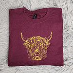 Maroon crewneck sweatshirt folded on a white faux fur rug, embroidered in gold thread with a shaggy Highland cow head with long curved horns