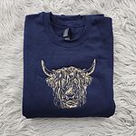Navy crewneck sweatshirt folded on a white faux fur rug, embroidered in light tan thread with a shaggy Highland cow head with long curved horns