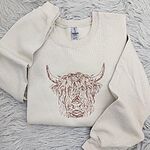 Sand crewneck sweatshirt laid on a gray faux fur rug, embroidered in copper thread with a shaggy Highland cow head with long curved horns