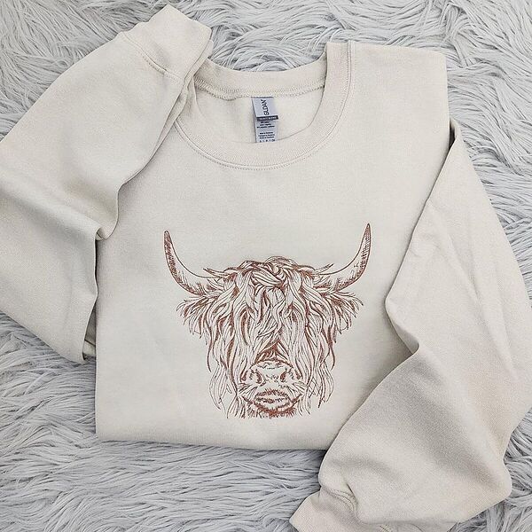 Sand crewneck sweatshirt laid on a gray faux fur rug, embroidered in copper thread with a shaggy Highland cow head with long curved horns