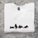White holiday cat crewneck with four black cats playing with Christmas lights, a red and green gift and red ornaments