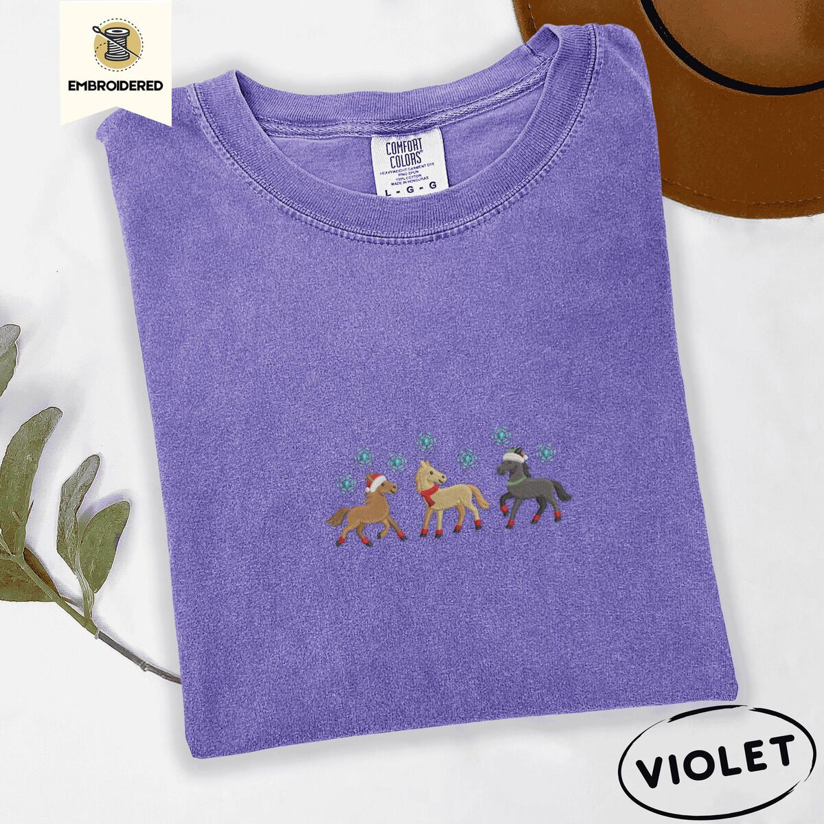 Horse Christmas Shirt – Violet Violet Comfort Colors T-shirt embroidered with three horses in Santa hats, a red scarf and red leg wraps trotting under turquoise snowflakes