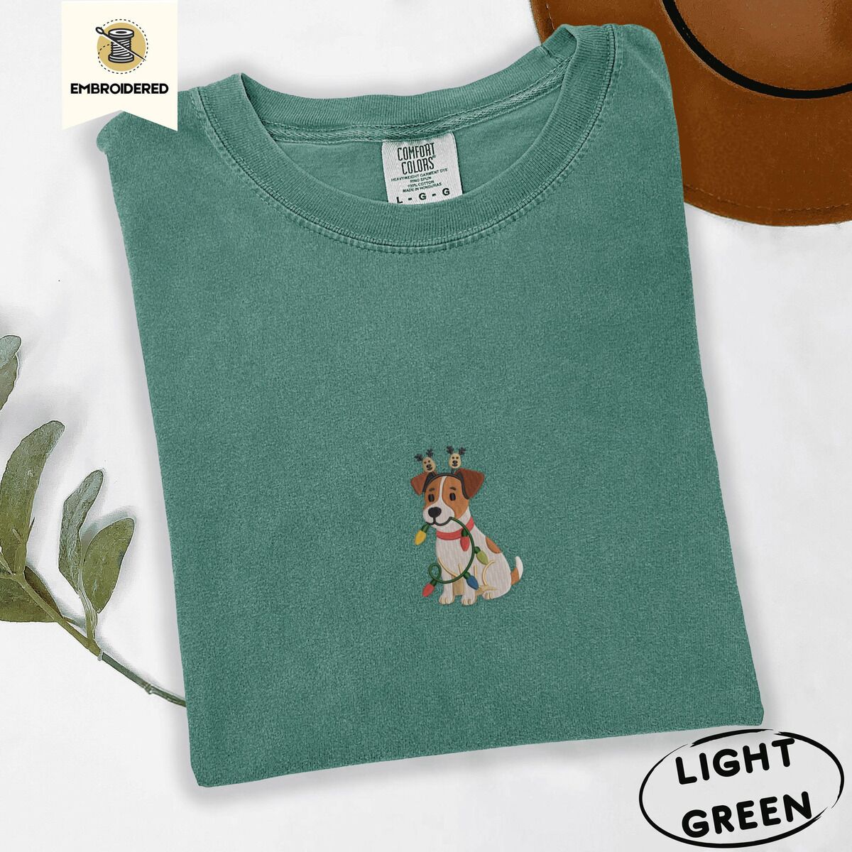 Jack Russell Christmas Lights Shirt – Light Green Light Green Comfort Colors T-shirt embroidered with a white and tan Jack Russell puppy in a reindeer headband chewing a string of Christmas lights