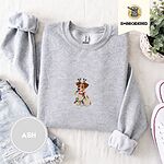 Ash crewneck sweatshirt folded with denim shorts, white sneakers and a potted plant, embroidered with a white and tan Jack Russell puppy in a reindeer headband chewing a string of Christmas lights