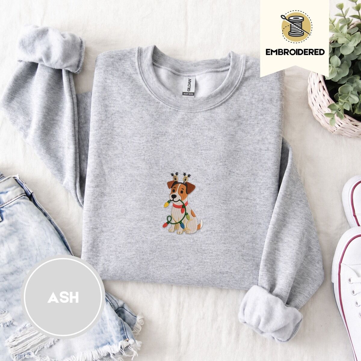Jack Russell Christmas Lights Sweatshirt – Ash Ash crewneck sweatshirt folded with denim shorts, white sneakers and a potted plant, embroidered with a white and tan Jack Russell puppy in a reindeer headband chewing a string of Christmas lights