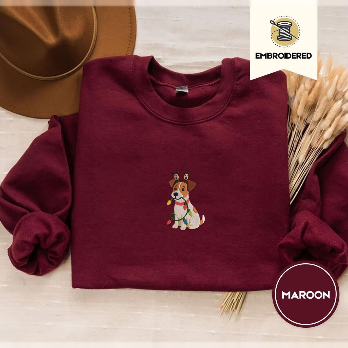 Jack Russell Christmas Lights Sweatshirt – Maroon Maroon crewneck sweatshirt with a brown felt hat and dried bunny tail grass, embroidered with a white and tan Jack Russell puppy in a reindeer headband chewing a string of Christmas lights