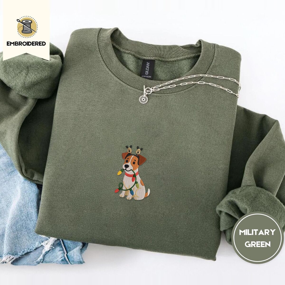 Jack Russell Christmas Lights Sweatshirt – Military Green Military Green crewneck sweatshirt folded with light jeans and a silver chain necklace, embroidered with a white and tan Jack Russell puppy in a reindeer headband chewing a string of Christmas lights
