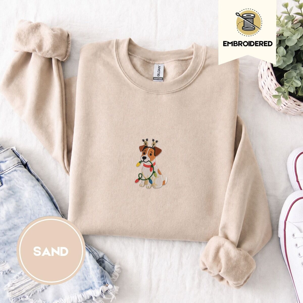 Jack Russell Christmas Lights Sweatshirt – Sand Sand crewneck sweatshirt folded with denim shorts, white sneakers and a potted plant, embroidered with a white and tan Jack Russell puppy in a reindeer headband chewing a string of Christmas lights