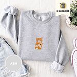 Ash crewneck sweatshirt folded with denim shorts, white sneakers and a potted plant, embroidered with two fluffy orange tabby kittens, one winking with a paw on the other's head