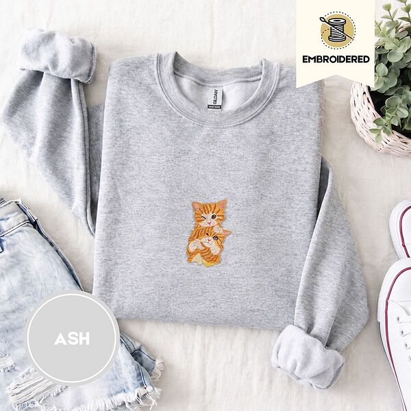 Ash crewneck sweatshirt folded with denim shorts, white sneakers and a potted plant, embroidered with two fluffy orange tabby kittens, one winking with a paw on the other's head