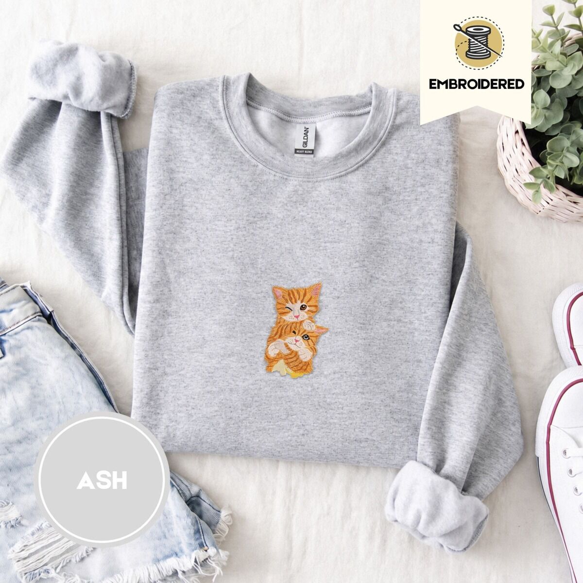 Kitten Sweatshirt – Ash Ash crewneck sweatshirt folded with denim shorts, white sneakers and a potted plant, embroidered with two fluffy orange tabby kittens, one winking with a paw on the other's head